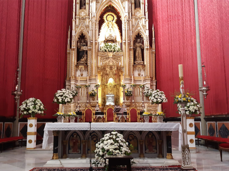 Interior de una iglesia con un altar dorado central, rodeado por flores blancas y rojas. La pared detrás es de color rojo brillante, con vitrales en el fondo. Hay sillas rojas alrededor del altar y un suelo de mármol blanco.