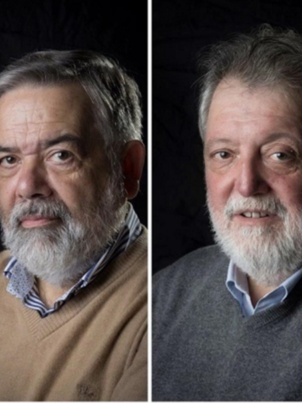 Dos hombres con barba y cabello gris, uno en suéter beige y otro en gris, frente a un fondo negro.