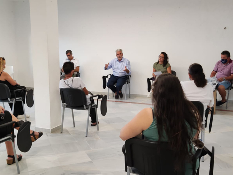En una sala de conferencias, un hombre en camisa azul habla a una audiencia compuesta por siete personas sentadas en sillas de diferentes colores. La imagen muestra una escena formal de comunicación o presentación, con el hablante en el centro y la audiencia atenta.