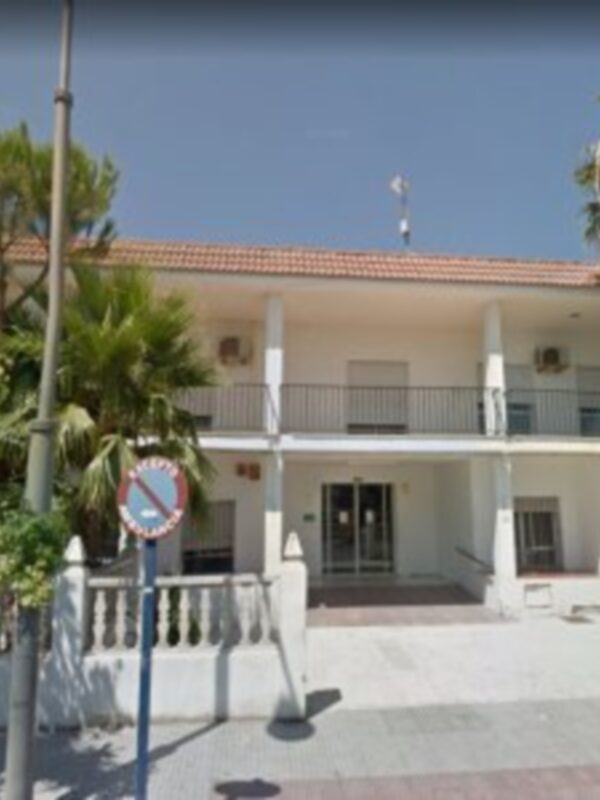 Fotografía de una casa blanca con balcones y ventanas, rodeada por árboles verdes. En la esquina inferior izquierda se puede observar un poste de luz con una señal circular roja y blanca. La imagen muestra una vista frontal del edificio, con cielo azul claro en el fondo.