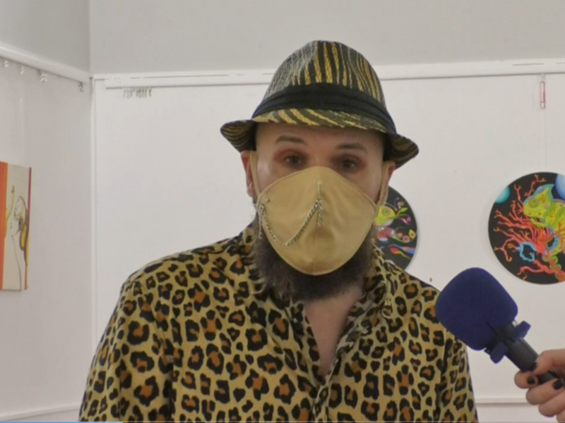 Person wearing a leopard print shirt, a hat, and a face mask, holding a microphone, in a room with colorful decorations.