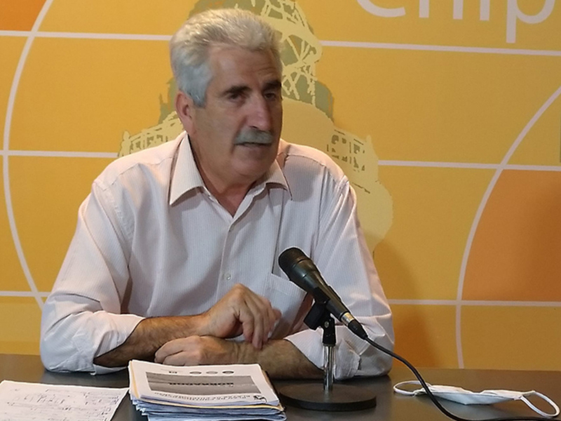 Persona hablando en rueda de prensa con micrófono y documentos en la mesa.