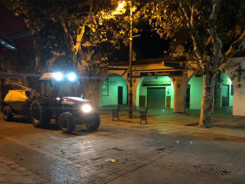 Un vehículo militar está estacionado en la noche frente a una estructura de madera con luces brillantes. La imagen muestra detalles urbanos nocturnos, incluyendo una calle con adoquines y un árbol en el lado derecho.