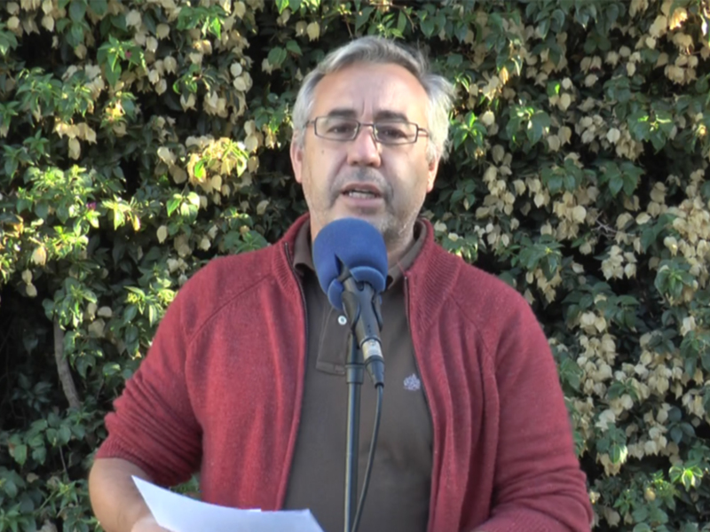 Un hombre con barba y gafas habla en un micrófono. Lleva una chaqueta roja y sostiene papeles en la mano izquierda. Fondo de vegetación verde con pequeñas flores amarillas.