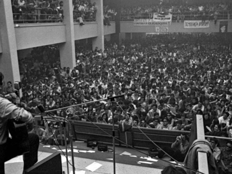 Un concierto en un estadio lleno de público, con un artista en el escenario tocando la guitarra y una multitud de personas escuchando.