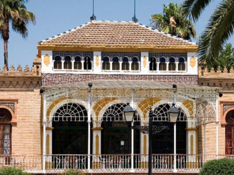 Un edificio colonial con tejas rojas, ventanas con marcos de madera y detalles dorados. En el frente hay un pórtico con columnas y una farola de metal. Al fondo, se pueden ver palmeras y un cielo azul claro.