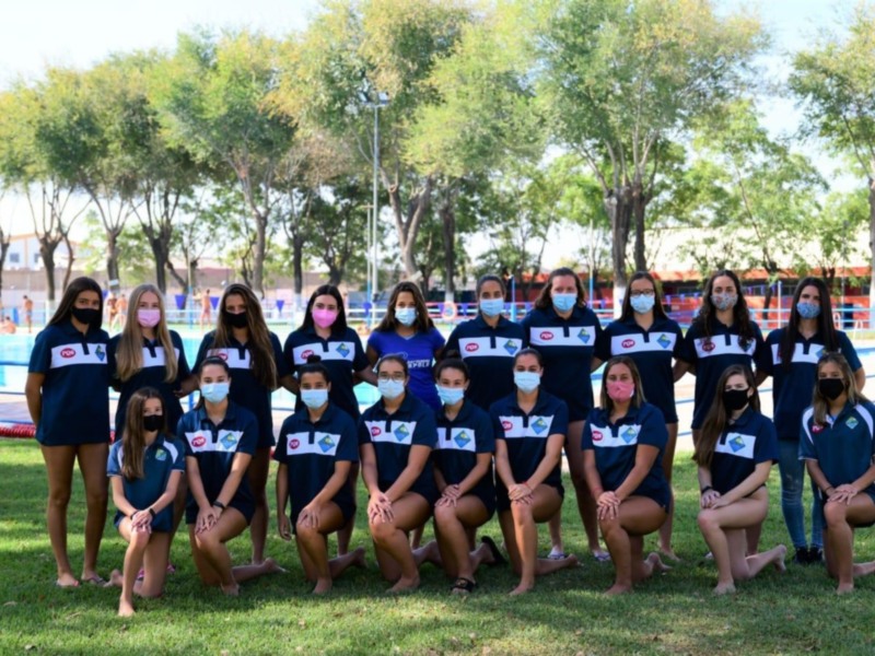 Equipo femenino de natación en una piscina, todos con máscaras y uniformes azules con logotipos, posando en una fila.