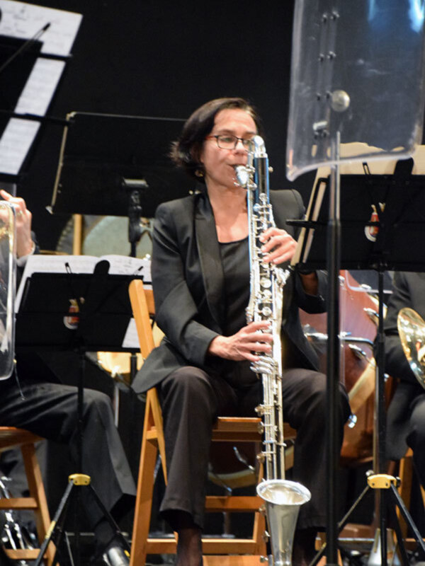 Una mujer toca el saxofón en un concierto de orquesta, con un piano y otros instrumentos musicales en el fondo.
