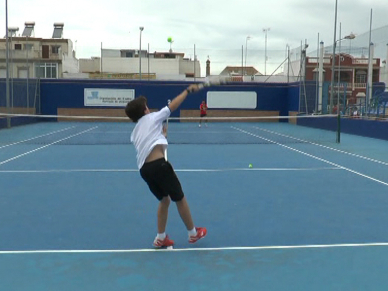 Un niño juega tenis en un campo azul con una línea blanca. Está de espaldas, preparándose para golpear la pelota que está en el aire. El fondo muestra edificios y una valla azul con un cartel blanco.