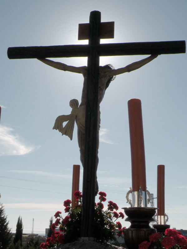 Crucifijo de madera con una figura en la cruz, rodeado de flores rojas y dos velas grandes. El cielo es claro y azul, con algunas nubes dispersas.