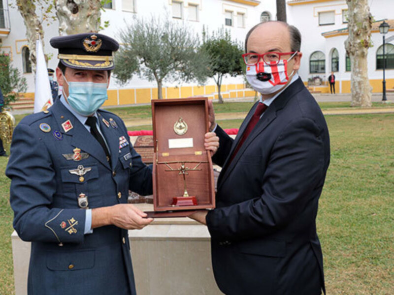 Dos hombres, uno en uniforme militar y otro en traje formal, sostienen una caja con un medalla de oro. El fondo muestra una escena al aire libre con árboles y una estructura blanca.