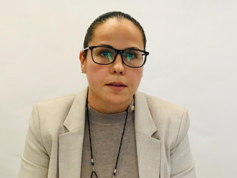 Mujer con gafas y chaqueta beige, expresando seriedad frente a un fondo blanco.