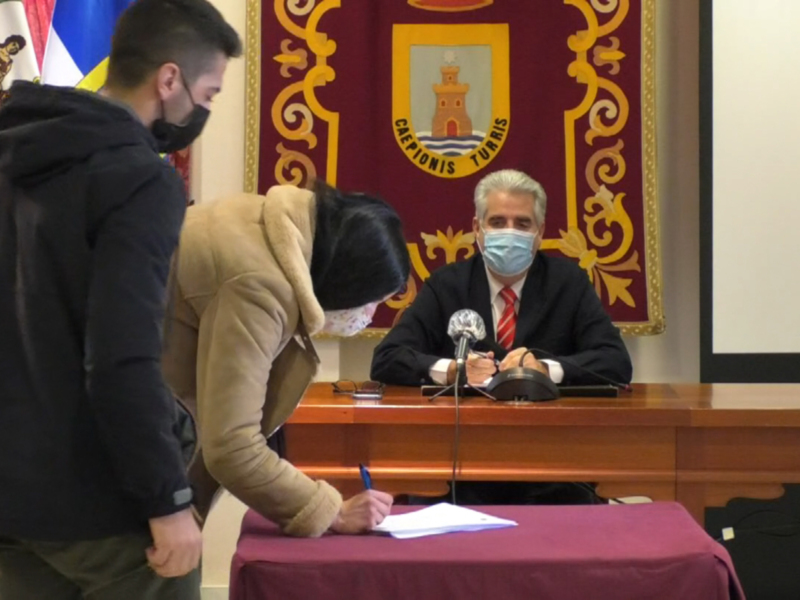 En una sala de audiencias, un hombre en traje y corbata se sienta detrás de una mesa con un micrófono. A su izquierda, dos individuos en sudaderas y máscaras se inclinan sobre una mesa para firmar un documento. En el fondo, banderas ondean y un escudo decorativo adorna la pared.
