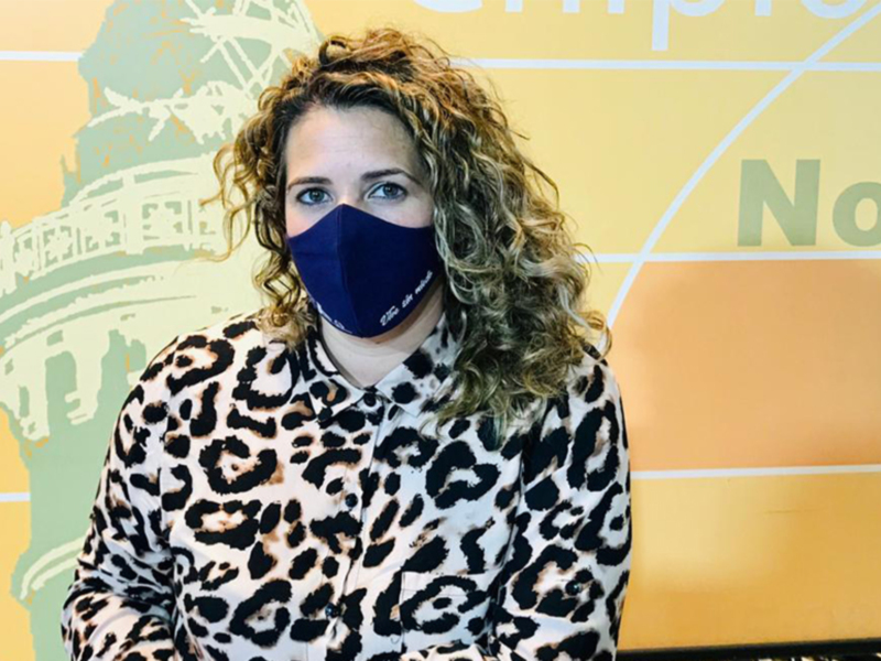 A una persona con cabello rizado y una máscara facial de color azul. El fondo es amarillo con un diseño gráfico que parece representar una estructura histórica. La persona está vestida con una chaqueta de manga larga que tiene un patrón leopardo.
