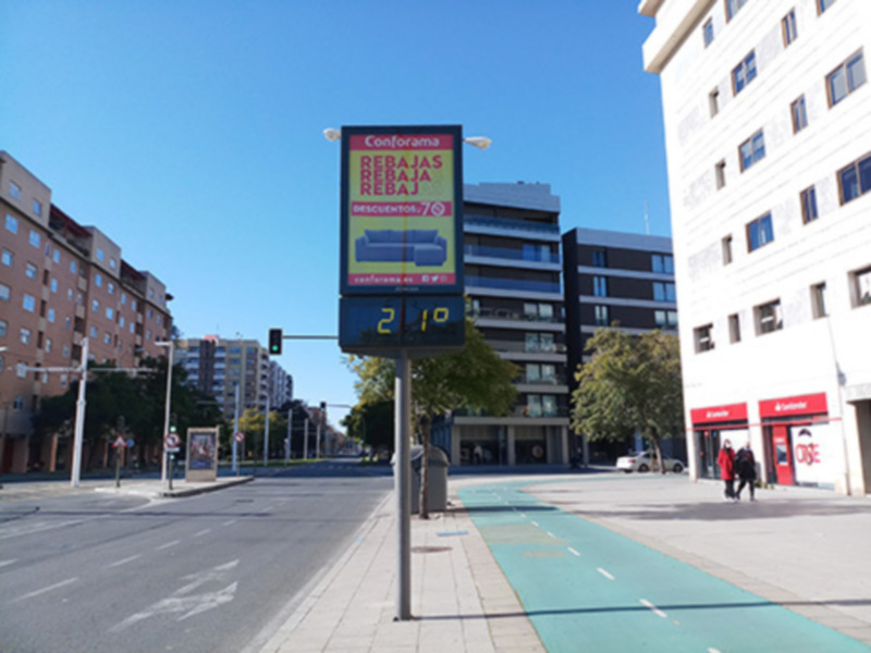 Se ve una calle con edificios a ambos lados, un semáforo en la esquina y una publicidad de Conforama con ofertas. La calle parece tranquila, con pocos vehículos y personas caminando.