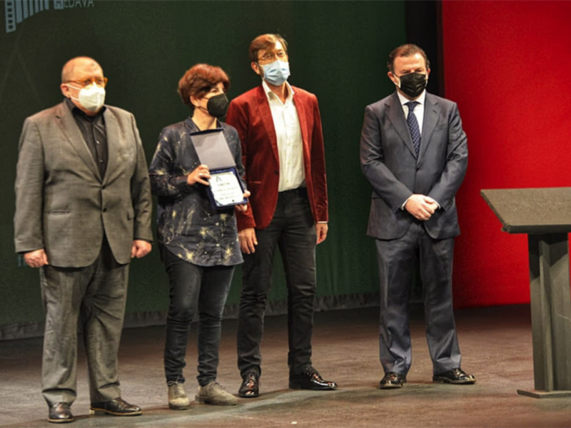 Una persona sostiene un premio mientras otros dos la observan en un escenario formal.