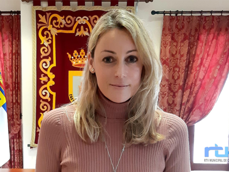 A una mujer con cabello rubio largo y ondulado, vestida con una camiseta de color rosa. Fondo: bandera y cortinas rojas.