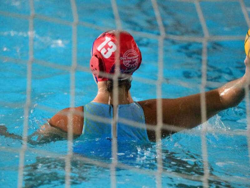 Un jugador de waterpolo en acción, con una pelota amarilla en la mano y un casco rojo con el número 13 y la frase 'Dos Hermanas juega limpio' en el fondo de la piscina azul.
