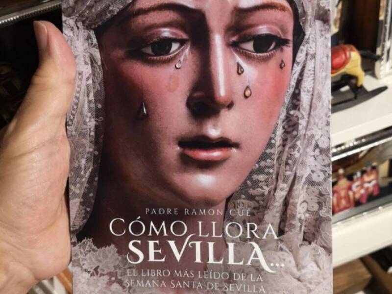 Un libro titulado "Cómo llora Sevilla" de Padre Ramón Cue, mostrado por una persona. La portada muestra una imagen de una estatua religiosa llorando, con gotas de agua en su rostro.