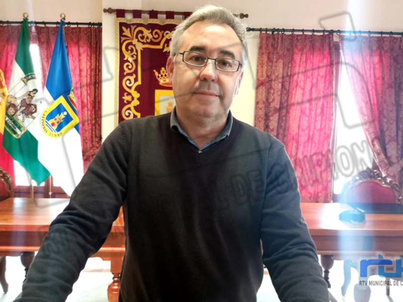 El pié de foto muestra a un hombre sentado en una mesa frente a varias banderas, incluyendo la de España y Andalucía. La imagen está tomada en un entorno formal, posiblemente una oficina o sala de conferencias.