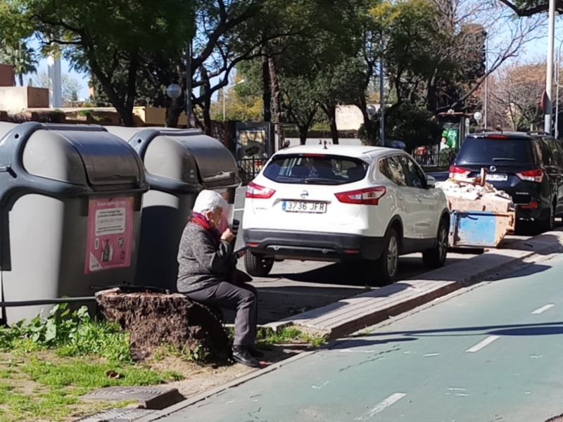 Un anciano se sienta en un tronco de árbol cerca de una calle con vehículos estacionados. Hay contenedores de basura y un cartel informativo en el fondo. La imagen muestra una escena urbana con vegetación y edificios a lo lejos.