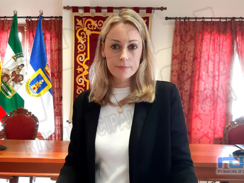 A una mujer con cabello rubio y vestida formalmente, posando frente a un escritorio. En el fondo se pueden ver banderas y cortinas rojas, lo que sugiere un ambiente oficial o institucional.