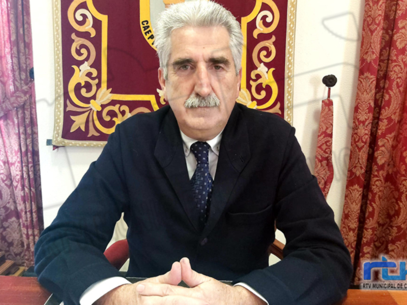 Un hombre con barba y bigote, vistiendo una chaqueta de traje y corbata, sentado frente a una bandera con símbolos geométricos. La imagen se encuentra en un entorno formal, posiblemente una oficina o sala de conferencias.