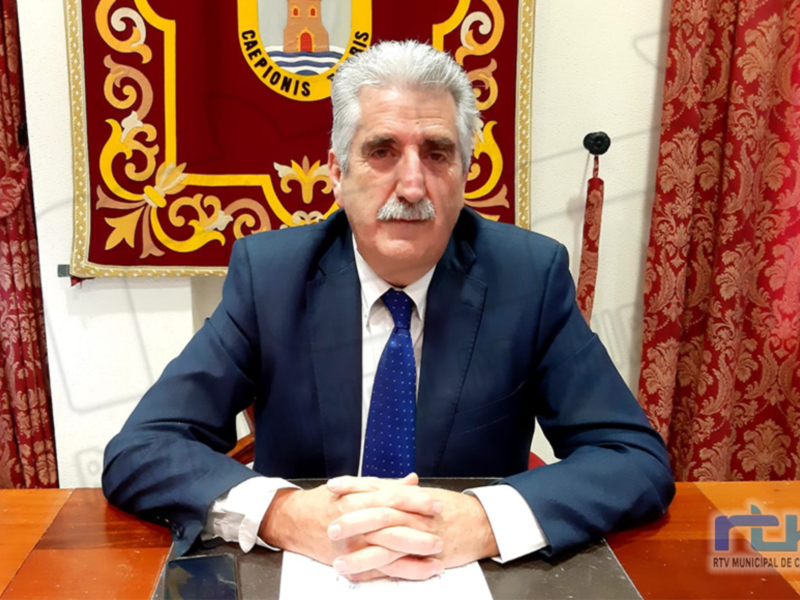 Persona sentada en un escritorio con un fondo de cortinas rojas y un escudo municipal, representando un entorno de oficina o gobierno.