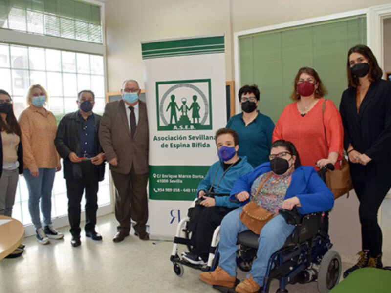 Una foto de una reunión en un espacio público. En el centro, una persona en silla de ruedas con una camiseta azul y pantalones azules, rodeada por otras personas vestidas de manera formal. Al fondo, un cartel verde y blanco con el logo de la Asociación Sevilla de Espina Bífida. Todos están mirando hacia la cámara, con expresiones serias y respetuosas.