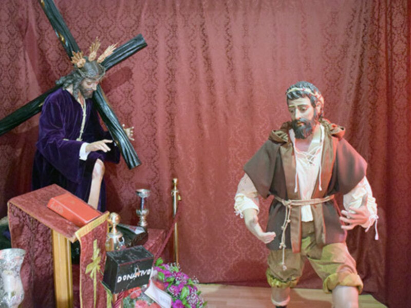 Figuras religiosas en una escena de la Pasión, con un personaje vestido con un hábito de sacerdote y otro con un traje de pobre, ambos con una cruz en el hombro. En el fondo, se puede ver un altar con una cruz y flores.