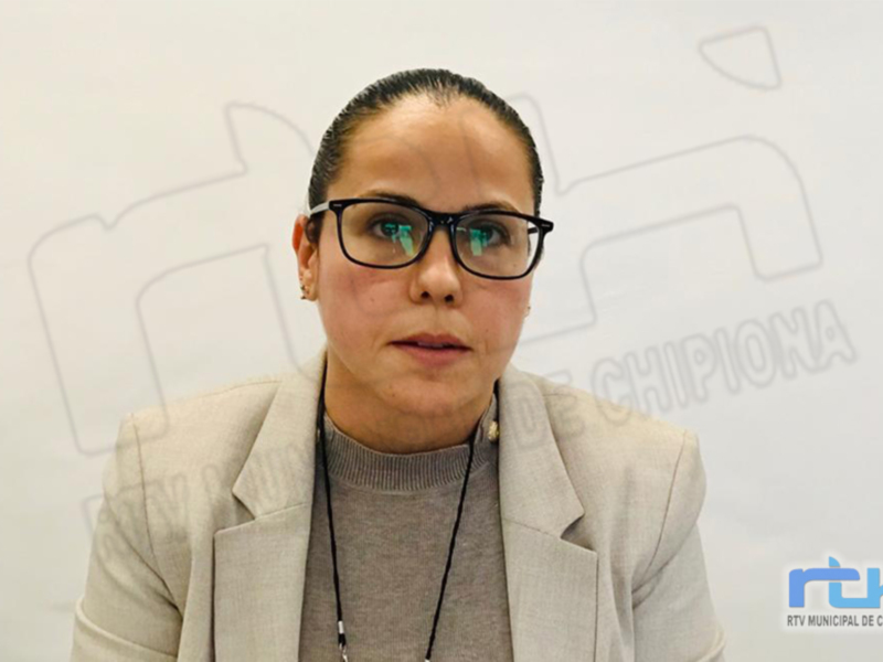 A una persona con cabello negro y rizado, peinado hacia atrás. Lleva gafas de montura oscura y una camisa gris con un chaleco beige. La persona está en un entorno interior con un fondo blanco y una marca visible en la parte superior derecha de la imagen.