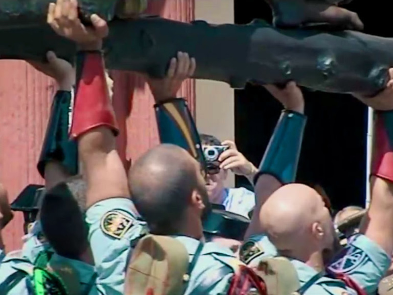 Una multitud de personas, vestidas con uniformes verdes y rojos, sostienen una gran estatua de un hombre en alto. La escena parece ser parte de una celebración o ceremonia importante, con algunos individuos tomando fotos. La imagen transmite un sentido de unidad y orgullo colectivo entre los participantes.
