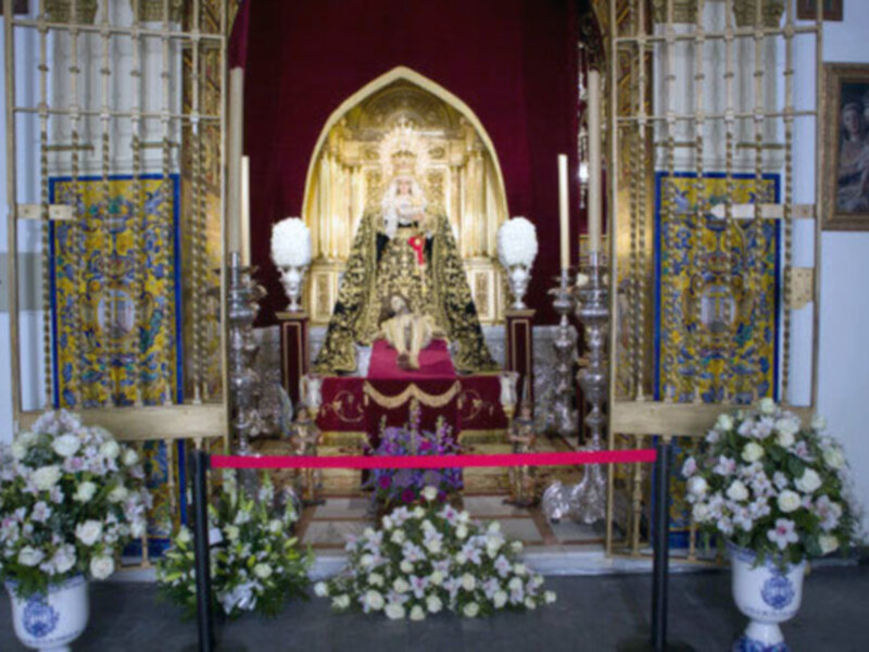 Una imagen de una estatua de oro en un altar, rodeada de flores blancas y doradas, con una profunda arquitectura de madera y un fondo rojo.
