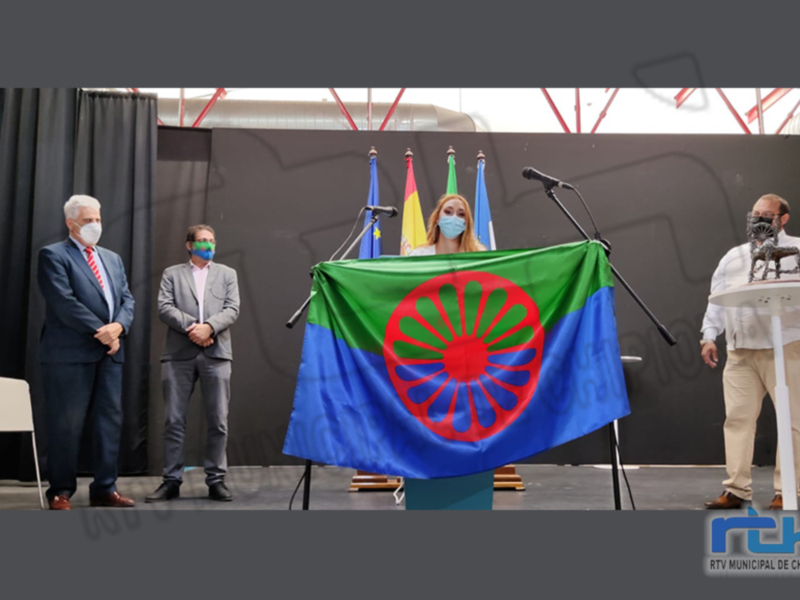 Event with three individuals standing behind microphones, one holding a flag with a red and blue wheel-like symbol on a green background.