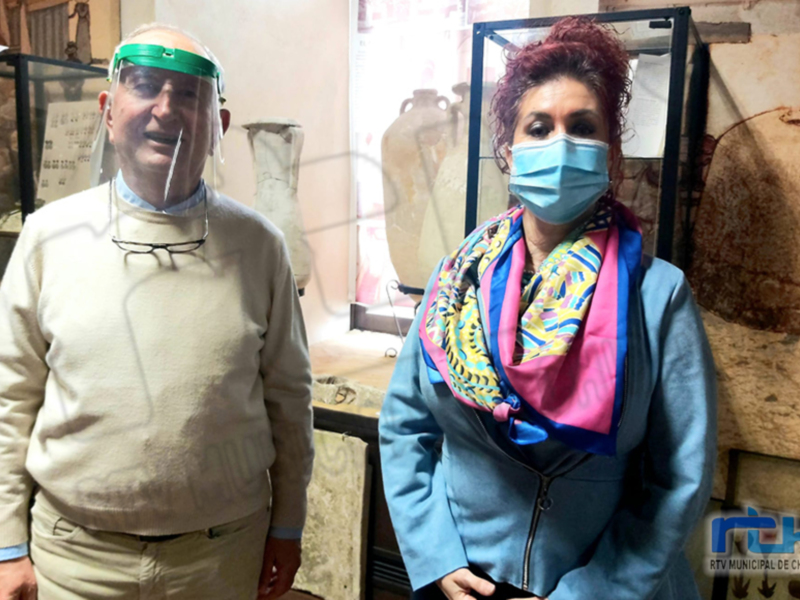 Dos personas con mascarillas y viseras en un museo, rodeadas de exposiciones de cerámica antigua.