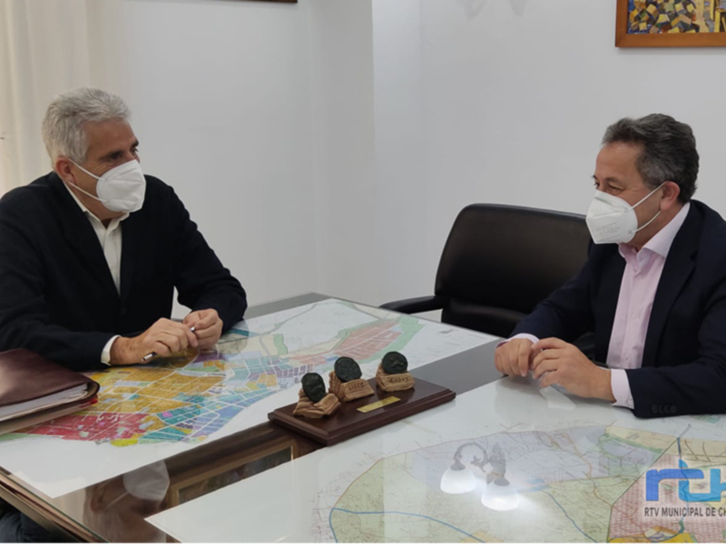 Dos hombres en una reunión en una oficina, discutiendo sobre un mapa y documentos, ambos con mascarillas y trajes formales.