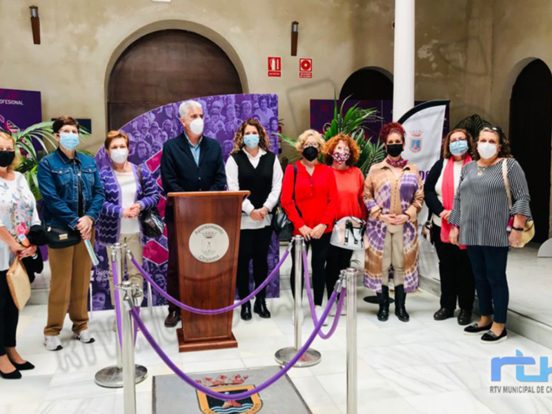 Una ceremonia formal en un edificio histórico, con una mesa de madera central y varios participantes vestidos formalmente. La imagen incluye elementos como una pancarta con texto, un mural de arte urbano y decoraciones florales.
