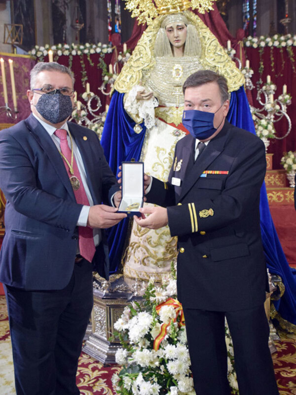 Un hombre en uniforme entrega una medalla a otra persona, ambos con máscaras faciales, en un ambiente festivo con una imagen religiosa en el fondo.
