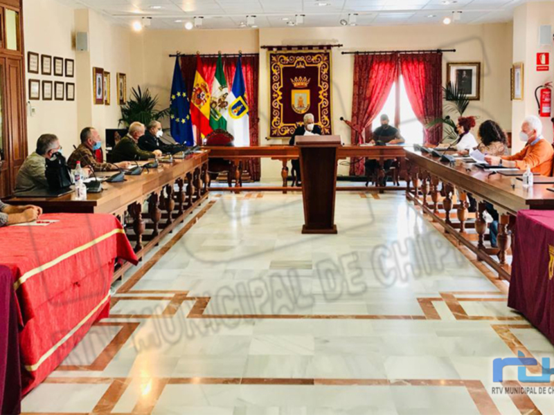 Sala de reuniones municipal con personas sentadas en mesas, banderas y emblemas en el fondo. El suelo es de mármol blanco y las mesas están cubiertas con mantas rojas. El ambiente es formal y ordenado, indicando una reunión oficial o administrativa.