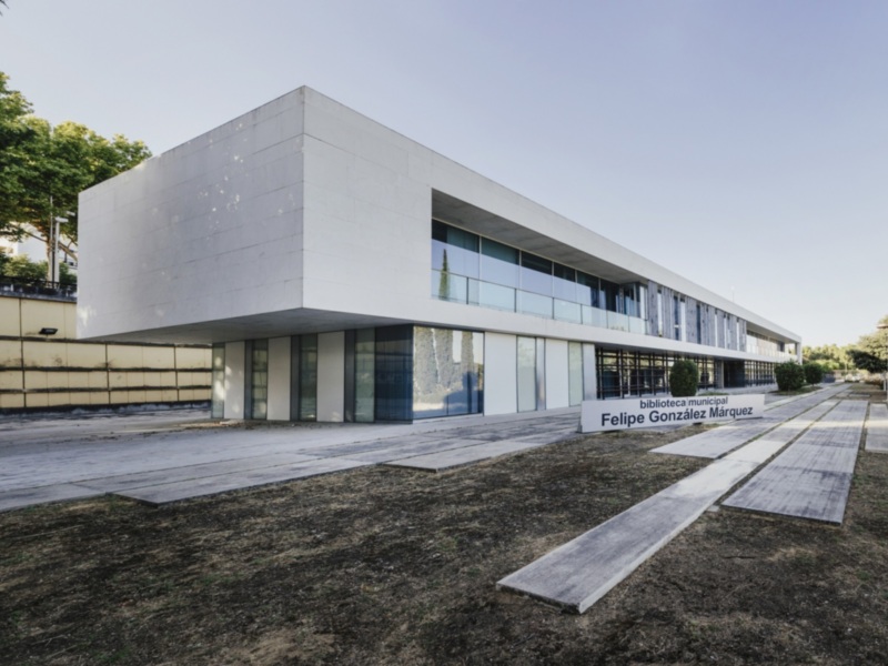 Biblioteca Municipal Felipe González Márquez, edificio moderno con fachada blanca y grandes ventanas. El área alrededor está rodeada de vegetación y el cielo es claro y azul.