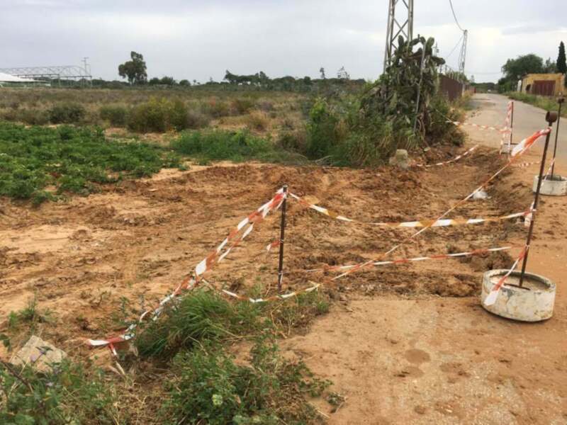 Un terreno desolado con un camino de tierra, barras de seguridad rojo y blanco, cactus en el fondo y un poste de electricidad.