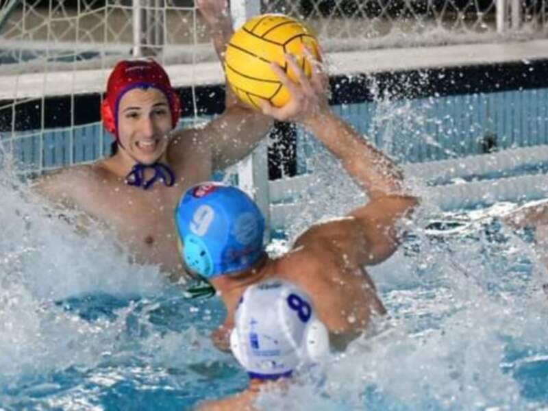 Jugadores de waterpolo en acción en una piscina, con el balón en el aire y los equipos de color azul y rojo.