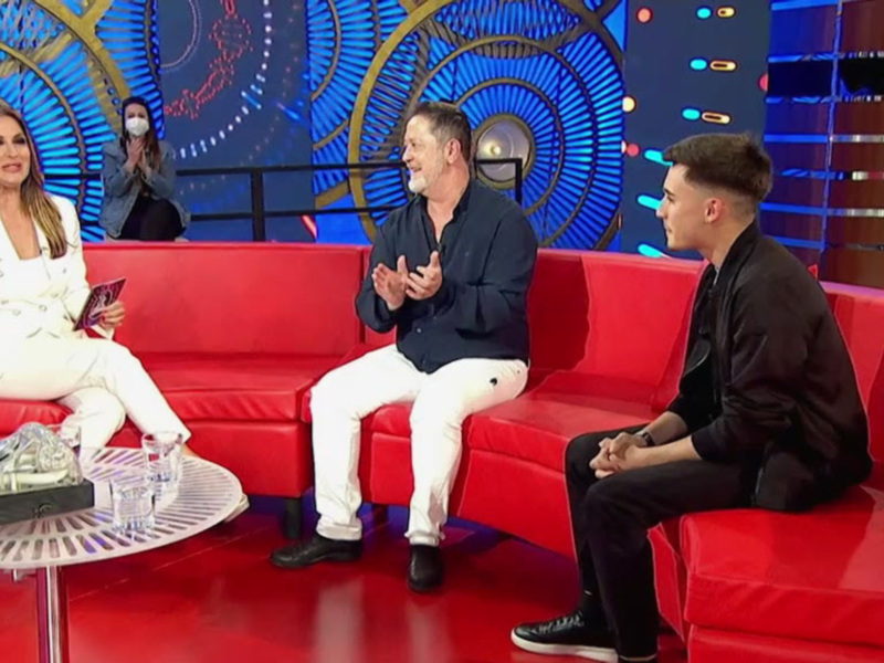 Tres personas sentadas en un sofá rojo en un programa de televisión, con una mesa de centro y un fondo con luces azules y doradas.
