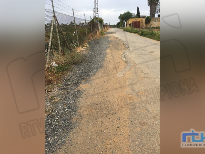 Un camino de tierra sin asfaltar con una cerca en el lado izquierdo. En la distancia, se observa un edificio y árboles. La imagen parece ser de una zona rural o suburbana.