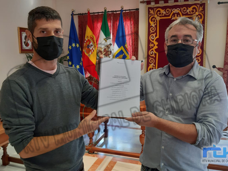 Dos hombres sostienen un documento, uno de ellos con una bandera española y otra europea detrás. La imagen parece ser de un evento oficial o formal, posiblemente relacionado con la administración local.
