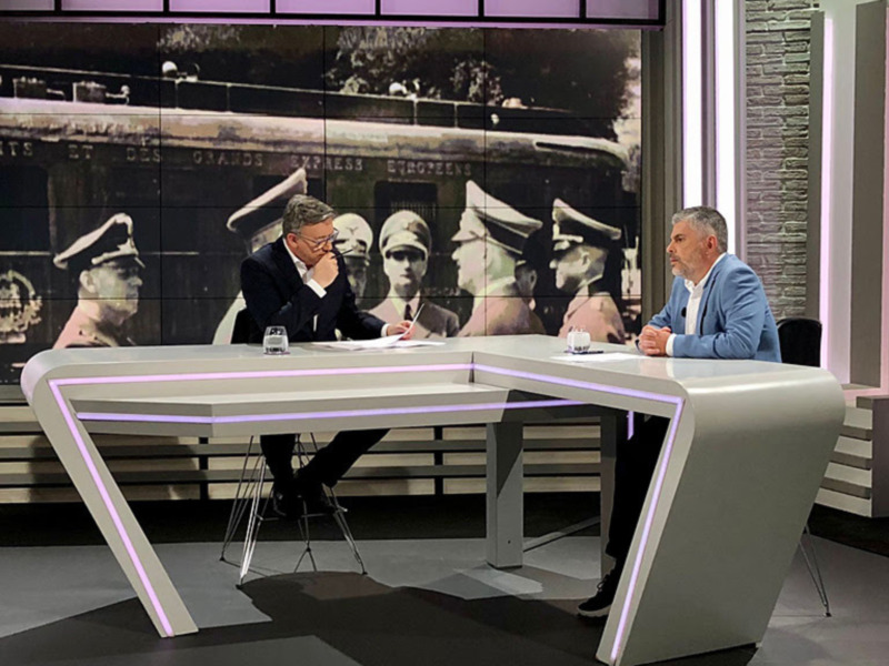 En un estudio de televisión moderno, dos hombres se sientan a una mesa blanca con iluminación de color púrpura. El fondo muestra imágenes antiguas de soldados con sombreros militares, sugiriendo un tema histórico o militar. La atmósfera es profesional y formal.