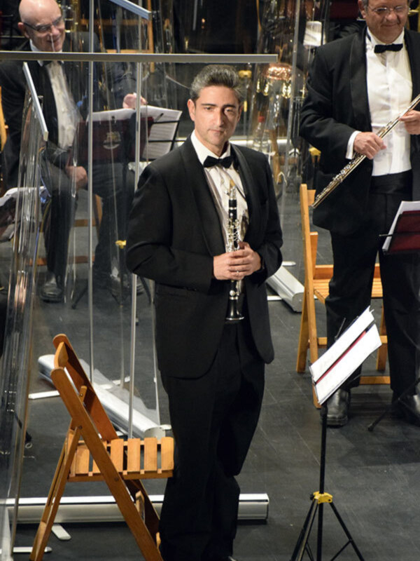 Un músico en traje negro y corbata sostiene un clarinete, preparándose para tocar. Detrás de él, otro músico también en traje negro y corbata, toca una flauta. Ambos están en un escenario con instrumentos de viento al fondo y una pizarra musical.