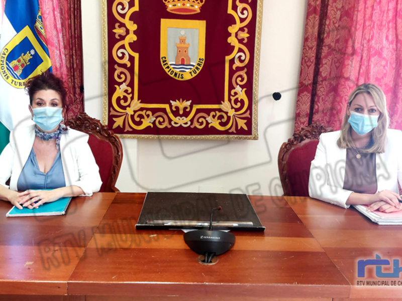 Dos mujeres sentadas en una mesa de madera frente a banderas y un escudo, ambas con mascarillas médicas.