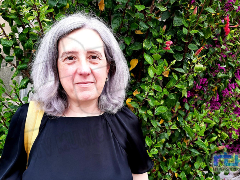 Una mujer con cabello gris, vestida de negro y con una mochila amarilla en el hombro, frente a un jardín de hojas verdes y flores rojas.