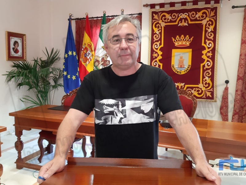 Un hombre con una camiseta negra y gafas de prescripción se encuentra sentado en un escritorio frente a una bandera española y la bandera de la Unión Europea. A su lado, hay una plantilla y un cuadro en el fondo.